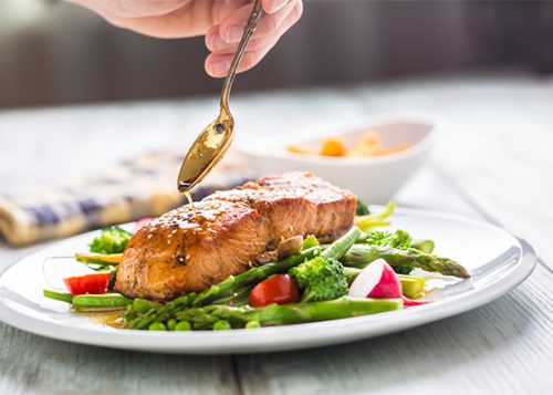 A salmon dinner plated with asparagus and other vegetables, a person spoons sauce onto it.