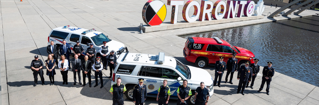 Three emergency vehicles parked in Nathan Phillips Square near the Toronto sign. Toronto Fire Services, Toronto Paramedic Services and Toronto Police services vehicles each with staff in dress uniforms standing in front of them.