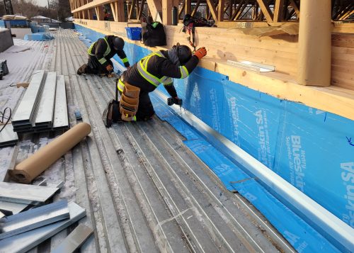 Roof installation in progress showing two construction workers kneeling on a corrugated metal deck and drilling into blue vapour barrier attached to the side of a wooden structure. Construction tools, metal beams and other materials are scattered around the work site, with some snow visible on the ground.