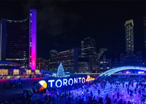 People skating in Nathan Phillips Square at night in front of Toronto City Hall and holiday glittering lights.