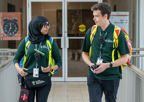 Two people dressed in green uniforms, safety vests and wearing intercoms talking to each other