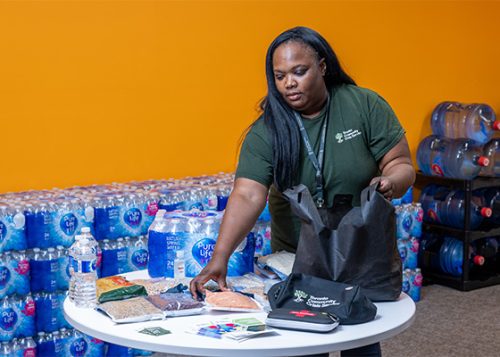 A woman packing a bag with food and supplies with several cases of bottled water behind her.