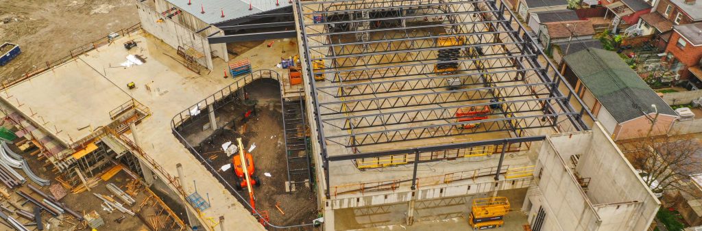 Close up aerial photograph of the construction site at Wallace Emersion Community Centre on May 5, 2024, which focuses on the Fieldhouse in the community centre roof, with steel scaffolding and beams, and the concrete lower levels. The community centre is rectangular with the front entrance curving outwards.