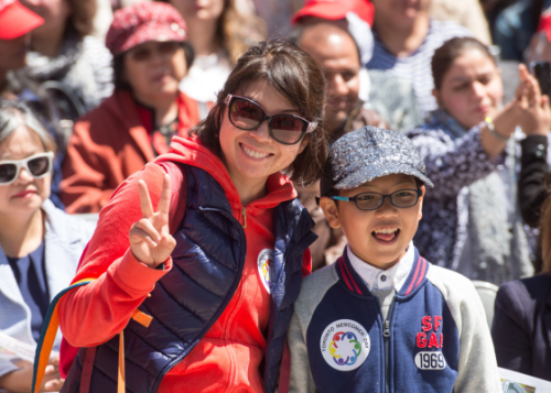 Adult wearing sunglasses and giving a peace sign standing next to a child with a crowd in the background.