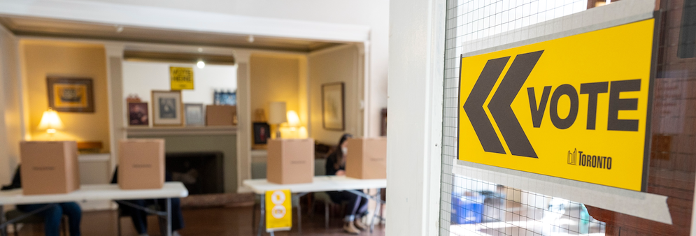 Entry of a voting place with a yellow vote sign directing voters into the voting place. In the background there are voting screens set up.