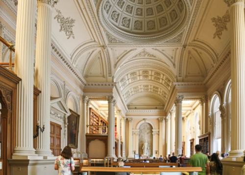 People walking through an ornate neo-classical room with Corinthian columns, vaulted ceiling and a niche with a sculpture
